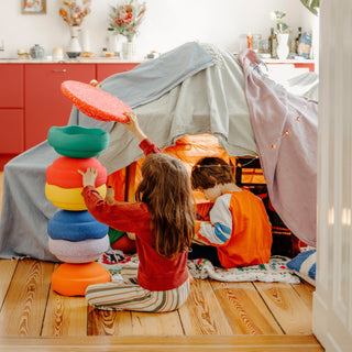 Two children playing in a cosy indoor play scene with a fabric tent and a stack of colorful Original Stapelstein pieces.
