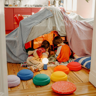 Two children play inside a fabric tent on a wooden floor, with colorful Original Stapelstein pieces arranged around them in a bright room.