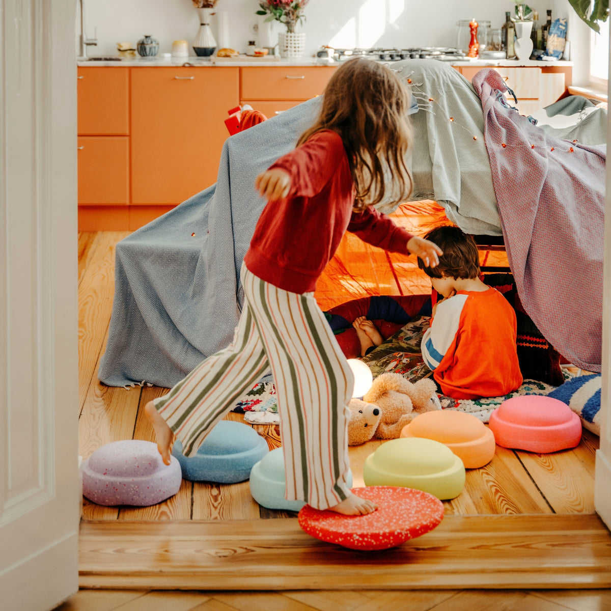 A little girl in a red sweatshirt and striped trousers stands barefoot on a red circular board while another child sits inside a play tent made from blankets, in a bright room with wooden floors and orange cabinets.
