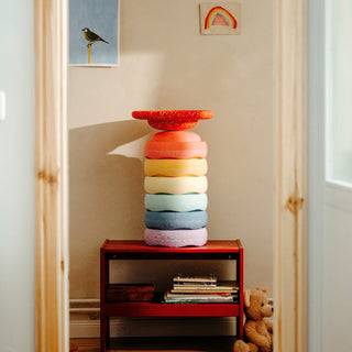 A colorful stack of round Original Staplestein pieces in red, orange, pink, yellow, light blue, dark blue, and lavender, with a red wobble board displayed on a small red shelf in a child's room.